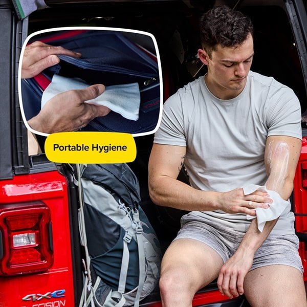 A man sits on the back of a red vehicle, cleaning his arm with a Skunky portable bathing sponge. An inset shows a hand pulling a Skunky wipe from a bag labeled "Portable Hygiene." A gray backpack sits nearby.