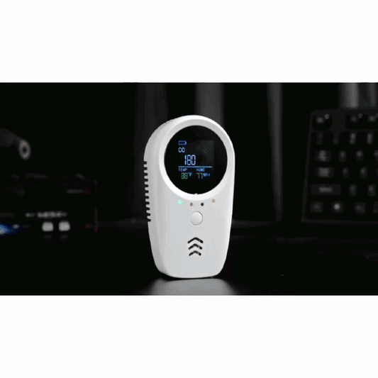 A close-up of the 5-in-1 Carbon Monoxide & Gas Detector with a circular display showing CO, temperature, and humidity readings, placed on a dark surface with a blurred keyboard and background.