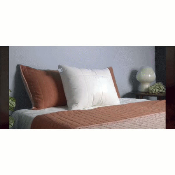 A neatly made bed features brown and white pillows, including the Adjustable Height Pillow for added comfort, topped with a brown quilted blanket. Beside the bed, a side table holds a white lamp and small plants against a gray wall.