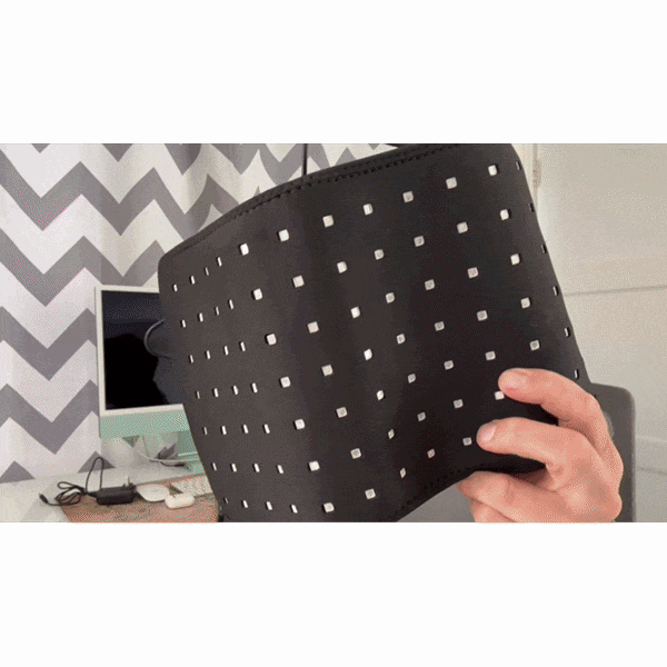 A hand holds up a black, square-patterned lampshade in front of a desk with a computer and chevron curtains—perfect to pair with Red Light Therapy Wrap for joint pain relief.