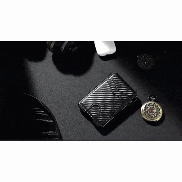 The Slim Compact RFID Wallet, crafted from black carbon fiber, is pictured open, securely holding cards. The minimalist wallet sits on a dark surface beside a watch and pocket watch, illuminated by dramatic lighting.