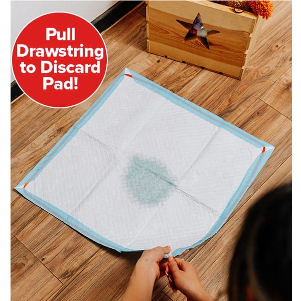 A person pulls the drawstring on a Drawstring Pee Pad (Includes 25 Pads), which has a wet spot in the center. Text in a red circle reads, "Pull Drawstring to Discard Pad!" The leak-proof pet pad is placed on a wooden floor near a box with a star cutout.