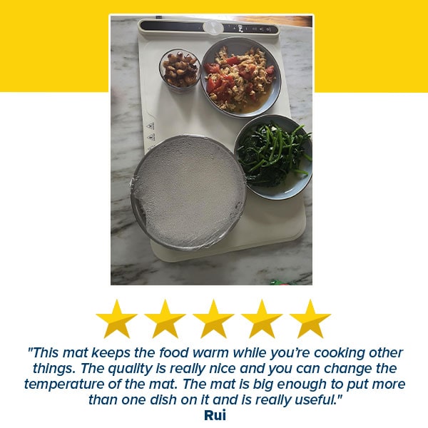 Overhead view of the Electric Food Warming Mat in white, crafted from food-grade silicone, warming three metal bowls of food, a small bowl of nuts, and a foil-covered plate; a five-star customer review is displayed below.