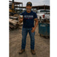 A smiling man in a Veteran shirt and cap stands on a construction site holding a small can of Hempvana Cocoa Butter pain relief cream. He is wearing jeans and work boots, with trucks, lumber, and a blue Jobox visible behind him.