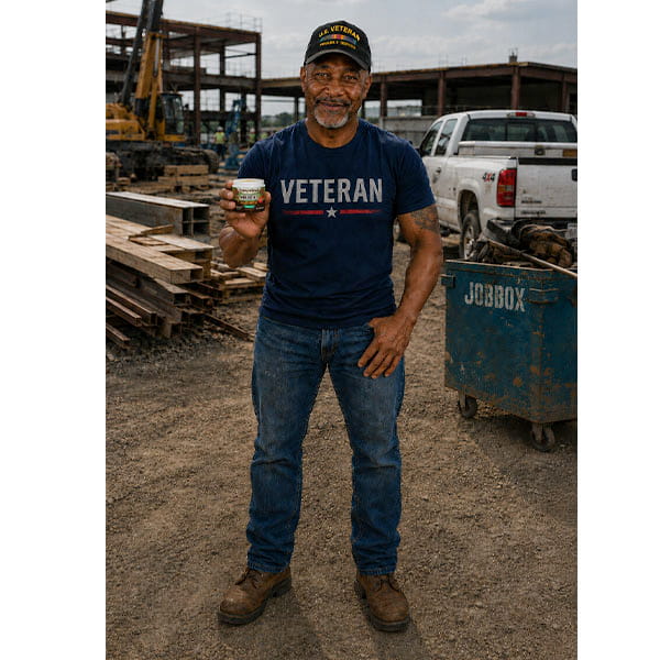 A smiling man in a Veteran shirt and cap stands on a construction site holding a small can of Hempvana Cocoa Butter pain relief cream. He is wearing jeans and work boots, with trucks, lumber, and a blue Jobox visible behind him.