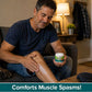 A man sits on a couch, smiling as he applies Hempvana Leg Cramps cream to his leg. He holds a container of the pain relief cream. Text at the bottom reads, Comforts Muscle Spasms! A lamp and coat rack are visible in the background.