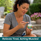 A woman in a gray t-shirt smiles while applying cream from a small container labeled Hempvana Leg Cramps, enriched with hemp seed oil. Text below reads: Relieves Tired, Aching Muscles! Outdoors with plants in the background.