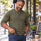 A man in a green long-sleeve shirt smiles as he slips the Slim Compact RFID Wallet into his jeans pocket, while a takeaway coffee cup rests on the outdoor café table next to him.