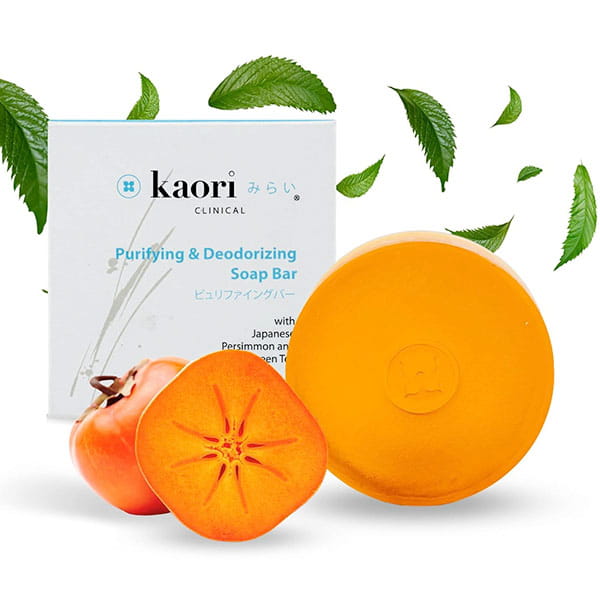A round Kaori Soap Bar for Old People Smell next to a sliced Japanese persimmon and its product box, with floating green leaves.