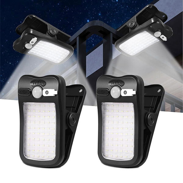Two black Clip On Outdoor Solar Lights (Pack of 2) with motion sensors are mounted on a wall at night, illuminating the area. Two more clip-on solar lights with adjustable panels are displayed in front for versatile outdoor lighting.