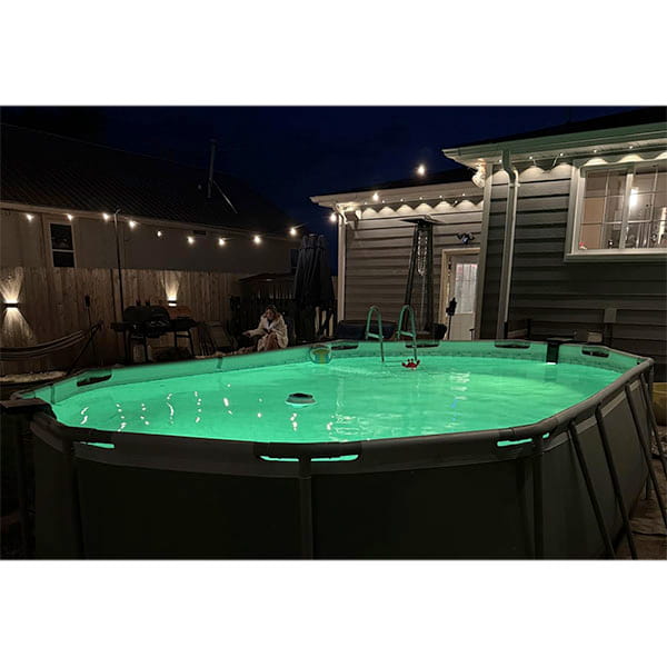 A backyard above-ground pool glows at night with Solar Pool Lights for Inground & Above Ground Pool, while string lights hang on the wooden fence and house. Cozy patio furniture and a hammock complete the relaxing scene.