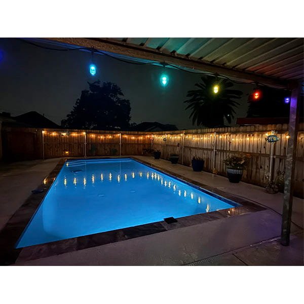 A backyard pool glows at night with Solar Pool Lights for Inground & Above Ground Pool, while colorful string lights hang from the patio roof and warm lights line the wooden fence. Potted plants enhance the inviting, illuminated poolside.