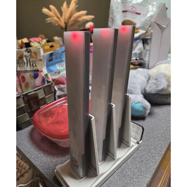 Three upright vacuums with red lights charge via a USB charging station on a gray countertop, lit by the 3-Pack Under Cabinet Lighting With Charging Station and surrounded by pet food, a stuffed toy, and a red-lidded container.