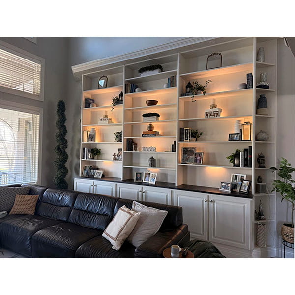 A living room with a dark sectional and pillows faces built-in white bookshelves filled with books, plants, and decor, all softly illuminated by the 3-Pack Under Cabinet Lighting With Charging Station.
