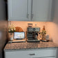 A granite kitchen countertop with a coffee machine, tablet displaying a building, decorative flowers, coffee bean jar, wooden tray, and white cabinets above illuminated by the 3-Pack Under Cabinet Lighting With Charging Station.