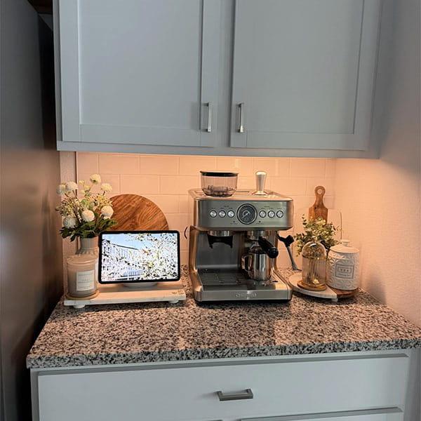 A granite kitchen countertop with a coffee machine, tablet displaying a building, decorative flowers, coffee bean jar, wooden tray, and white cabinets above illuminated by the 3-Pack Under Cabinet Lighting With Charging Station.